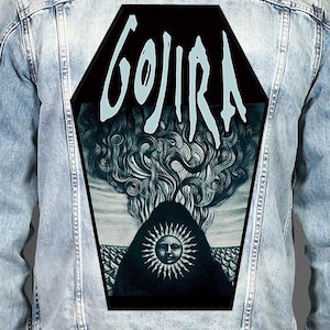 Peut inclure: Une veste en jean bleu clair avec un grand écusson dorsal en forme de cercueil. L'écusson affiche le mot GOJIRA en lettres blanches stylisées au-dessus d'une illustration sombre d'un soleil à visage humain sur des vagues et de la fumée.