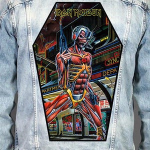 May include: A light blue denim jacket featuring a large, coffin-shaped back patch. The patch displays a colourful illustration of a cyborg character holding a futuristic weapon, with the text "IRON MAIDEN" printed in yellow at the top.