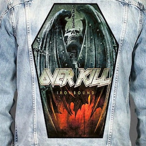 Può includere: Una giacca di jeans azzurra con una grande toppa posteriore a forma di bara. La toppa mostra un teschio alato con catene, il testo OVERKILL e la parola IRONBOUND in giallo, su uno sfondo scuro dai toni arancione fuoco e nero.