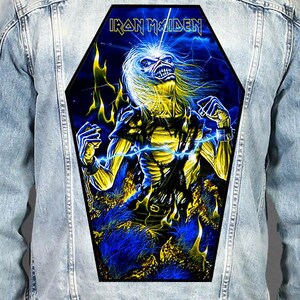Può includere: Una giacca di jeans azzurra con una grande toppa posteriore a forma di bara. La toppa mostra un'immagine della mascotte degli Iron Maiden, Eddie, in giallo e blu, circondata da fiamme e fulmini, con il nome della band Iron Maiden in alto.