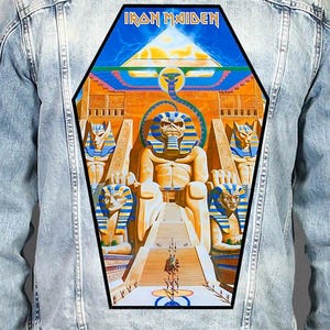 Op de afbeelding: Een lichtblauw spijkerjack met een grote, doodskistvormige rugpatch met een Egyptisch thema. De afbeelding toont een centrale faraofiguur, sfinxen en de tekst Iron Maiden in een vet, gestileerd lettertype aan de bovenzijde.
