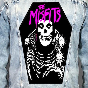 May include: A light blue denim jacket featuring a large, coffin-shaped black patch on the back. The patch displays a white skeleton graphic with spiked shoulder details and the text THE MISFITS in bright pink, dripping-style lettering.