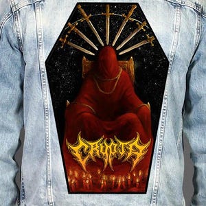 May include: A light blue denim jacket featuring a large, coffin-shaped back patch. The patch displays a hooded figure in a red robe sitting on a throne, surrounded by seven swords, a starry background, and the yellow text CRYPTA above red candles.