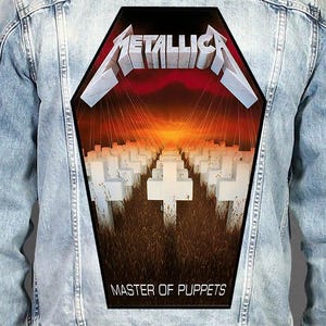 Può includere: Una giacca di jeans azzurra con una grande toppa posteriore a forma di bara. La toppa mostra il logo Metallica in bianco, un campo di croci bianche nell'erba alta e la scritta Master of Puppets su un cielo al tramonto rosso e arancione.