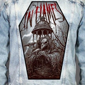 May include: A light blue denim jacket featuring a large, coffin-shaped back patch. The patch displays a dark, detailed illustration of a hooded figure with a bird-like mask standing under a gazebo, with the text IN FLAMES printed in red above.