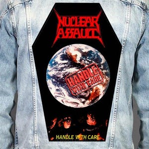Könnte beinhalten: Eine hellblaue Jeansjacke mit einem großen, sargförmigen Rückenaufnäher. Der Aufnäher zeigt den roten Schriftzug "NUCLEAR ASSAULT" über einem Bild der Erde mit einem roten "HANDLE WITH CARE"-Stempel und dem Text "HANDLE WITH CARE" unten.