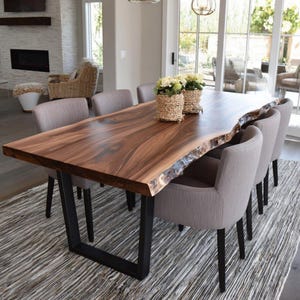 May include: A substantial dining table with a live edge, crafted from dark brown wood. It features black metal legs and is surrounded by six grey upholstered chairs. Two woven baskets with flowers are placed on the table. The table is approximately 240 cm long.