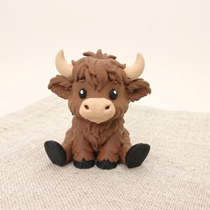 May include: A small, sculpted figurine of a Highland cow with dark brown, textured fur, a light beige muzzle, and curved, light-colored horns. The creature has large, dark eyes and black hooves, depicted in a seated position with a shaggy appearance.