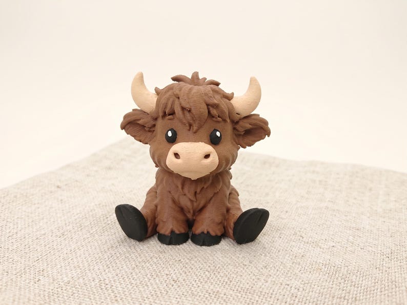 May include: A small, sculpted figurine of a Highland cow with dark brown, textured fur, a light beige muzzle, and curved, light-colored horns. The creature has large, dark eyes and black hooves, depicted in a seated position with a shaggy appearance.