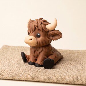 May include: A small, sculpted figurine of a brown Highland cow calf with a shaggy texture, light tan horns, a pale muzzle, and black hooves. The calf is depicted in a sitting position with its front legs extended forward and its head turned slightly.