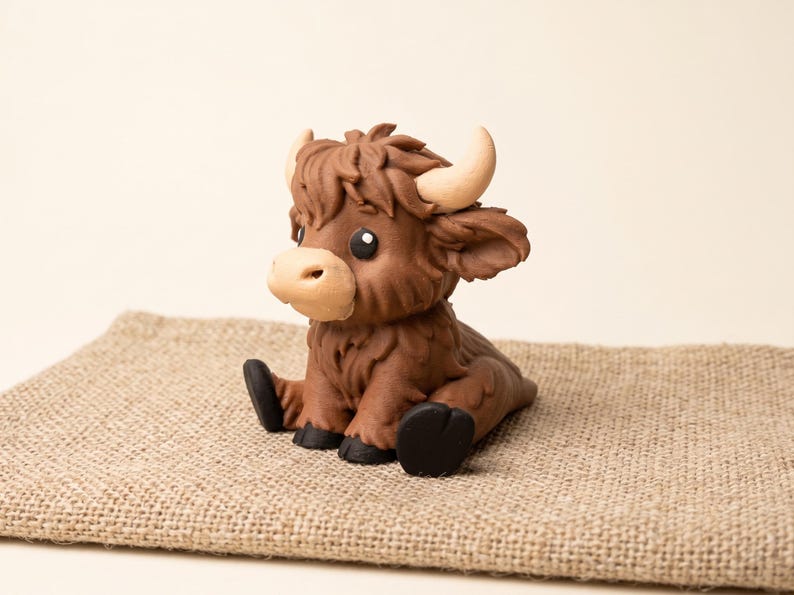 May include: A small, sculpted figurine of a brown Highland cow calf with a shaggy texture, light tan horns, a pale muzzle, and black hooves. The calf is depicted in a sitting position with its front legs extended forward and its head turned slightly.