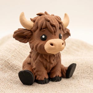 May include: A small, brown Highland cow figurine with a textured, shaggy coat, light tan horns, and a light tan muzzle. The figure has large, dark eyes with white highlights and black hooves, depicted in a seated position with a forward-facing gaze.