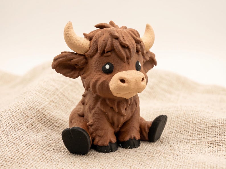 May include: A small, brown Highland cow figurine with a textured, shaggy coat, light tan horns, and a light tan muzzle. The figure has large, dark eyes with white highlights and black hooves, depicted in a seated position with a forward-facing gaze.