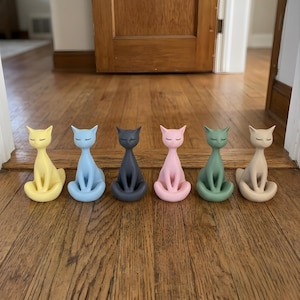 May include: A set of six cat figurines in a seated position with closed eyes. The cats are colored in yellow, light blue, dark gray, light pink, sage green, and beige. Each figurine features a smooth finish and a slender, upright posture.