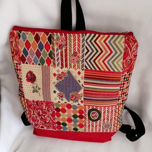 Puede incluir: Mochila de patchwork multicolor con base y ribete rojos, correas y asa negras. El diseño de patchwork incluye patrones florales, geométricos y a rayas. La bolsa está hecha con una variedad de telas.