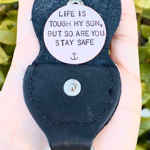 May include: A black leather keychain with a round copper charm that reads "Life is tough my son, but so are you. Stay safe." with an anchor symbol below the text.