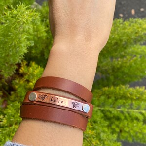 May include: A brown leather wrap bracelet with a copper plate engraved with the name "La'ie" and a small turtle design. The bracelet has two silver-toned metal accents.