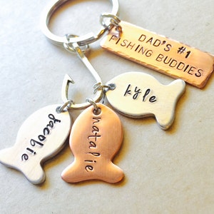 May include: A silver key chain with a copper tag that reads "Dad's #1 Fishing Buddies". There are three fish-shaped tags with the names "Kyle", "Natalie", and "Darcy" engraved on them.
