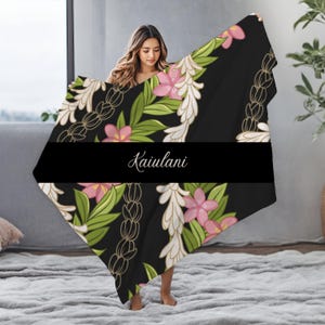 May include: A black and white floral patterned throw blanket with pink flowers and green leaves. The blanket has the word "Kaiulani" written in white text on a black background.