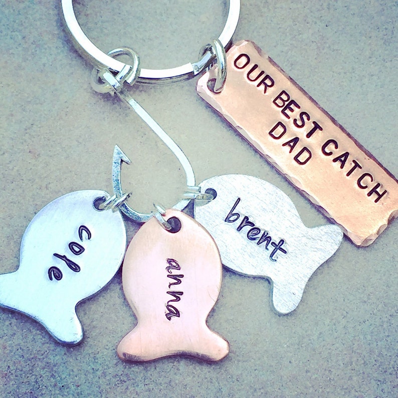 May include: A silver key chain with a fish hook charm and three fish charms. The fish charms are engraved with the names "Anna", "Cole", and "Brent". A copper tag is attached to the key chain and reads "Our Best Catch Dad".
