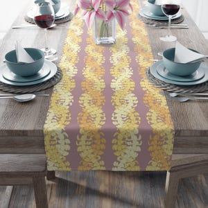 May include: A mauve table runner featuring a repeating pattern of yellow and gold floral designs. The runner is set on a wooden table, with place settings, wine glasses, and a vase of pink lilies. The table setting is for a formal dinner.
