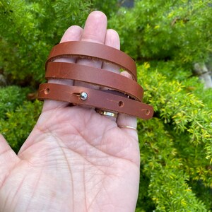 May include: A brown leather wrap bracelet with a silver metal accent. The bracelet is wrapped around a hand.