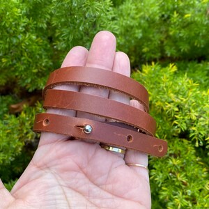 May include: A brown leather wrap bracelet with a silver metal clasp. The bracelet is wrapped around a hand, showing the multiple layers of leather.