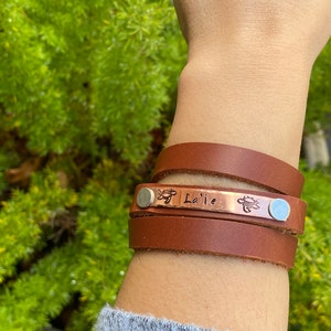 May include: A brown leather wrap bracelet with a copper plate engraved with the name "La'ie" and two turtle designs. The bracelet is worn on a person's wrist.