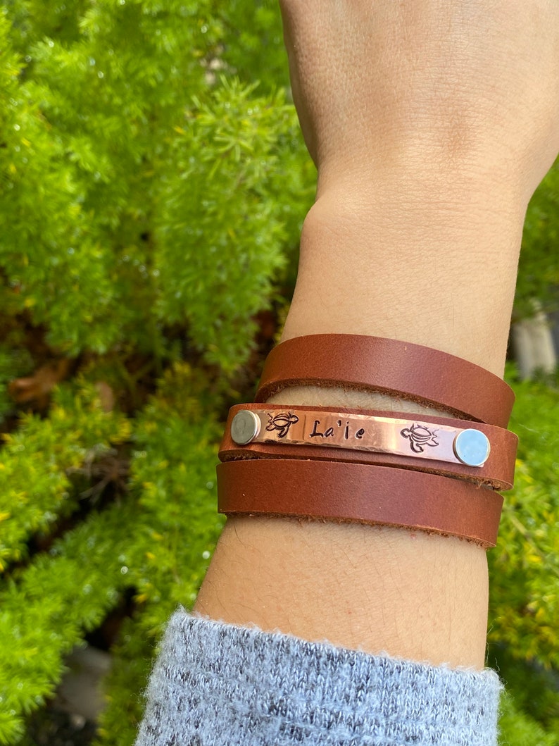 May include: A brown leather wrap bracelet with a copper plate engraved with the name "La'ie" and two turtle designs. The bracelet is worn on a person's wrist.