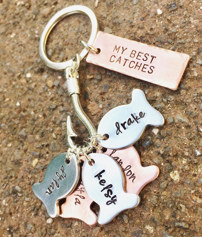 May include: Silver and copper fish-shaped charms with names on them, attached to a silver hook and a silver key ring. A copper tag with the text "MY BEST CATCHES" is also attached to the key ring.