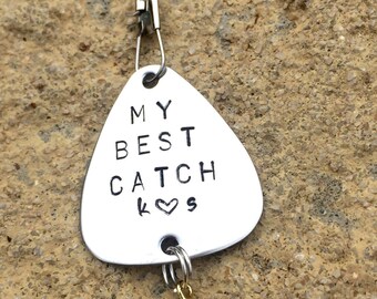 Op de afbeelding: Zilveren vislokaas met een hart en de tekst "My Best Catch" erop. Het lokaas is bevestigd aan een gouden haak.