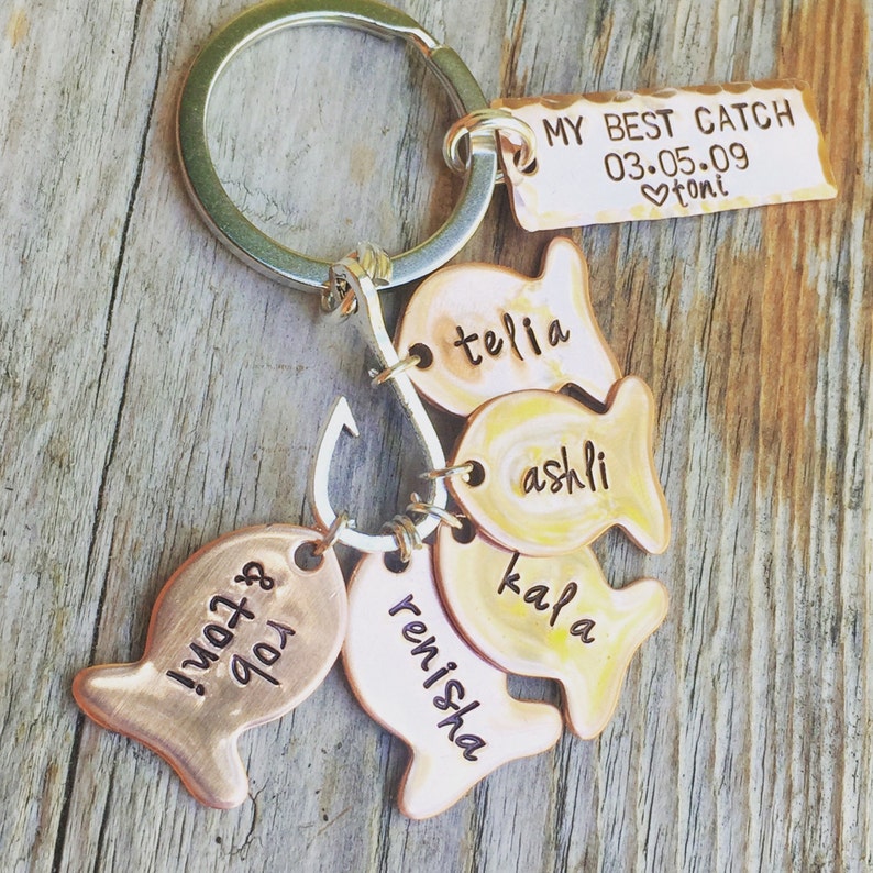 May include: A copper-toned keychain with a silver key ring and a hook. The keychain has five fish-shaped charms with the names "Telia", "Ashli", "Kala", "Renisha", and ""My"  "Tot"!" engraved on them. A rectangular charm with the text "My Best Catch 03.05.09 &hearts;Toni" is also attached to the keychain.