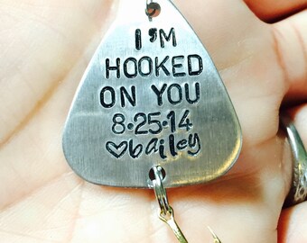Op de afbeelding: Een zilveren vislood met een zwarte inscriptie die "I'm Hooked On You 8-25-14 ♥ bailey" luidt. Het lood heeft een gouden ketting en twee gouden haken.