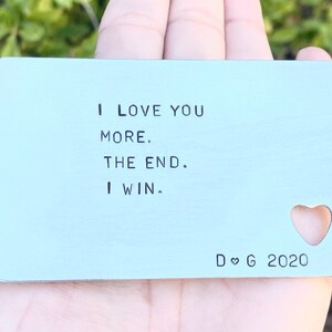 May include: A silver metal card with the text "I love you more. The end. I win." and a heart cutout. The card also has the text "D ♥ G 2020" at the bottom.