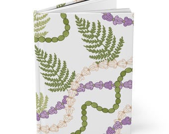 Può includere: Un diario con copertina rigida con sfondo bianco e un motivo ripetuto di fronde di felce verdi, fili di perline verdi e viola e fili di perline color crema. Il diario è aperto per rivelare pagine bianche vuote.
