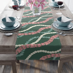 May include: A rectangular table runner featuring a dark green background with a repeating pattern of wavy, intertwined floral garlands in shades of pink, cream, and light blue. The runner is centred on a wooden table with place settings.