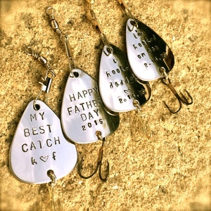 May include: Five silver fishing lures with personalized messages. The lures have gold hooks and are arranged on a textured brown surface. The messages on the lures read: "My Best Catch k &hearts; f", "Happy Father Day 2015", "I hooked daddy 2014.15", "I'm hooked on you 2.14.15".