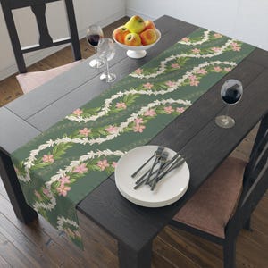 Pode incluir: Um corredor de mesa verde com um padrão floral branco e rosa. O corredor é feito de tecido e tem uma textura tecida. Está colocado sobre uma mesa de madeira escura com duas cadeiras e uma tigela de fruta.