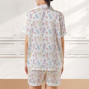 May include: White pajama set with a pastel bunny and bow pattern. The short-sleeved top and shorts feature a repeating design of bunnies in blue, yellow, pink, and purple, along with matching bows. The sleepwear set is made of a soft, lightweight fabric.