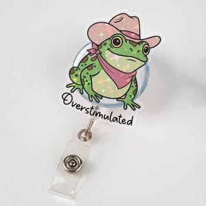 Puede incluir: Un carrete de insignia con una rana de dibujos animados que lleva un sombrero de vaquero y un pañuelo rosa. La rana es verde con manchas marrones y el texto "Overstimulated" está escrito debajo.