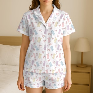 May include: White pajama set with a pastel bunny and bow pattern. The short-sleeved top has a notched collar and a chest pocket. The matching shorts complete the sleepwear ensemble. The pajamas are made of a soft, lightweight fabric.