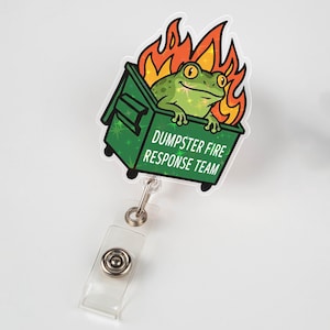 May include: A badge reel featuring a green frog in a green dumpster engulfed in orange flames. The text "DUMPSTER FIRE RESPONSE TEAM" is printed on the dumpster. The reel has a clear plastic strap and a metal clip.
