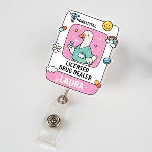 May include: A white and pink badge reel with the text "HONKSPITAL" and "LICENSED DRUG DEALER LAURA". The badge features a cartoon duck wearing scrubs and holding a pill bottle, with a rainbow, pills, and a smiley face.