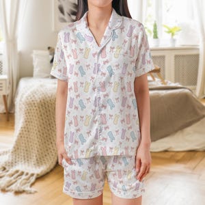 May include: White pajama set with a pastel bunny and bow pattern. The set includes a short-sleeved button-down shirt and matching shorts. The pajamas have a relaxed fit and are made of a soft, lightweight fabric.