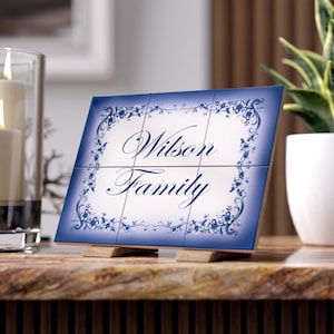 Peut inclure: Une plaque décorative rectangulaire composée de six carreaux présentant un centre blanc avec le texte bleu foncé Wilson Family. La bordure affiche un motif de vigne florale bleu foncé qui s'estompe sur le fond blanc, posée sur des supports en bois.