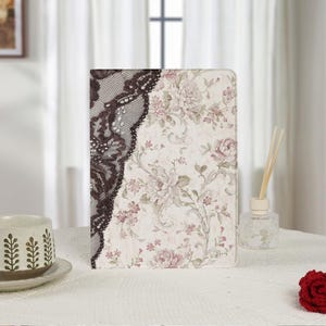 Puede incluir: Una funda rectangular para tableta con fondo color crema y un suave estampado floral en rosa y verde. El lado izquierdo de la funda está superpuesto con un diseño de encaje marrón oscuro intrincado que contrasta con el delicado estampado.