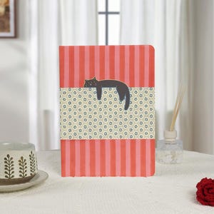 Op de afbeelding: Een decoratief notitieboek met een rood-roze gestreepte achtergrond en een bloemmotiefband. Een donkergrijze kattenillustratie rust op de bloemenband. Het notitieboek ligt op een wit oppervlak.