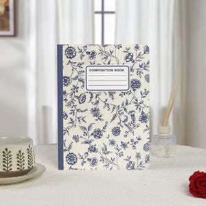 Puede incluir: Un cuaderno de composición con una cubierta crema con un estampado floral azul. El lomo es azul y la cubierta tiene una etiqueta que dice "COMPOSITION BOOK". El cuaderno está sobre una superficie blanca.