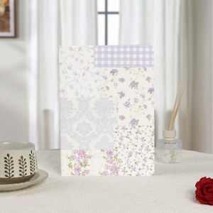 Könnte beinhalten: Ein rechteckiges Notizbuch mit Patchwork-Design. Das Notizbuch zeigt verschiedene Blumen- und Damastmuster in Weiß und Lavendel, mit einem lavendelfarbenen Vichy-Muster oben. Das Notizbuch liegt auf einer weißen Tischdecke.