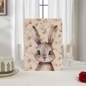 Puede incluir: Panel decorativo rectangular con una ilustración de acuarela de un conejo con orejas largas. El conejo está sobre un fondo beige con un estampado floral de rosas rosas. Una taza y un difusor están en el fondo.